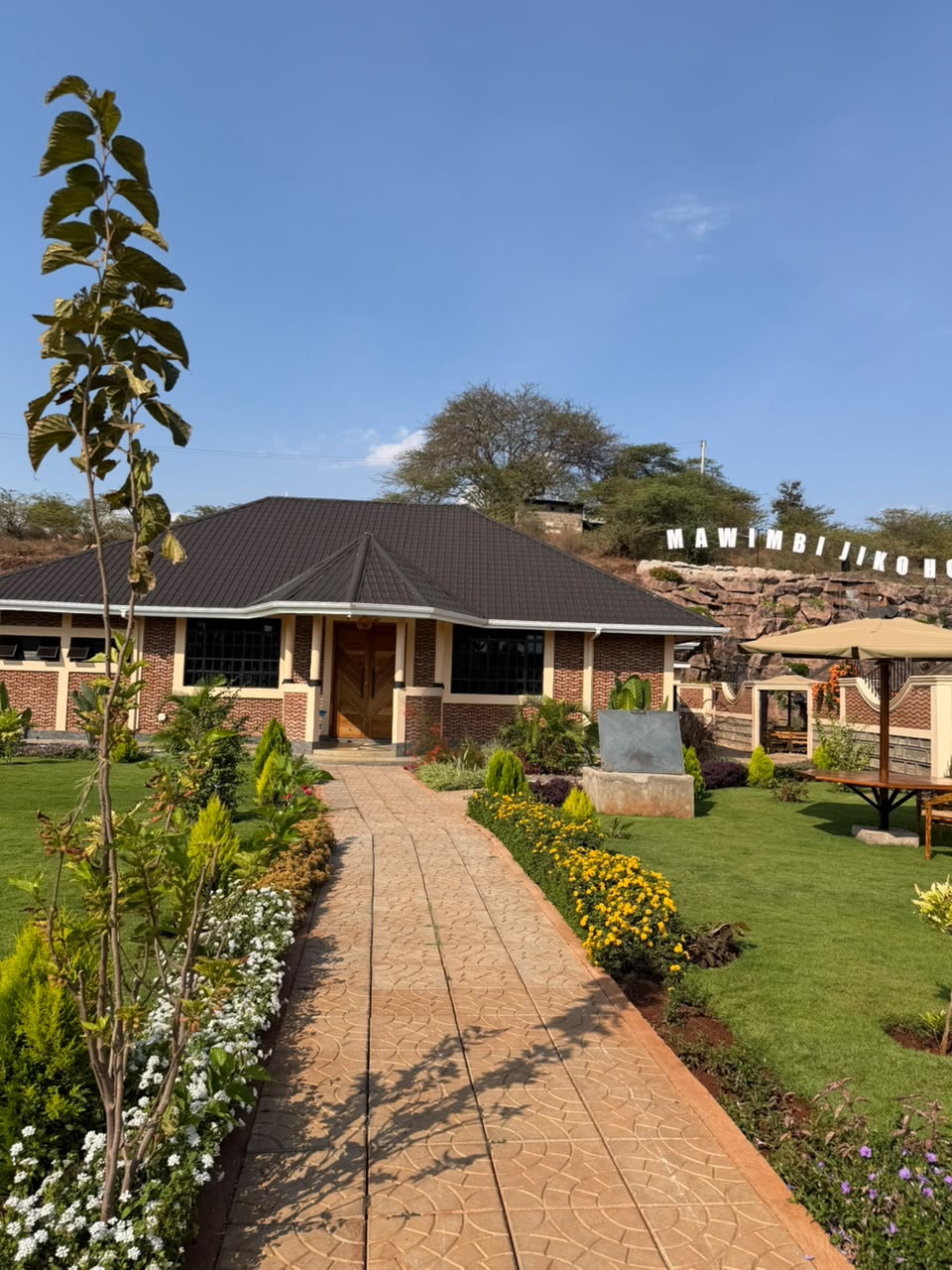 Paved walkway lined with flowering borders leading to the carved timber doors of the Mawimbi Jiko conference hall