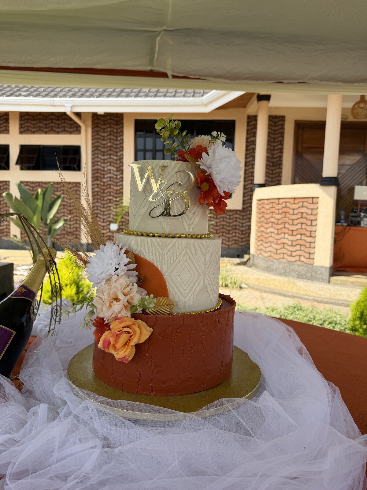 Three-tier birthday cake in terracotta and cream with gold monogram and fresh florals, set up outside the Mawimbi Jiko wave-brick building
