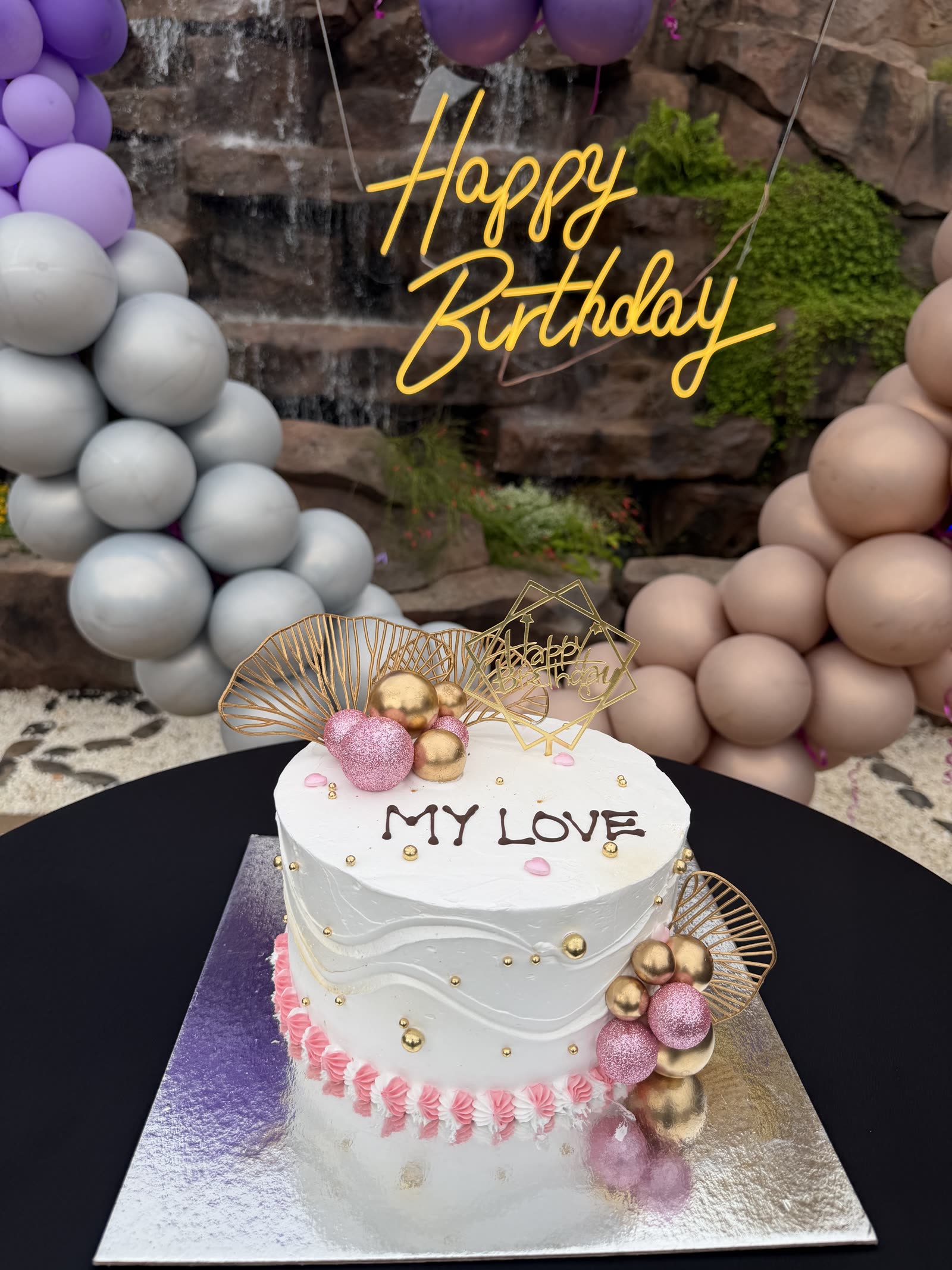 Birthday cake reading 'My Love' set against the neon Happy Birthday sign and balloon garlands at Mawimbi Jiko