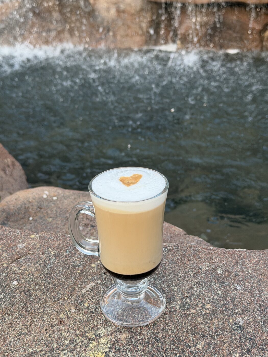 Mocha in a glass mug topped with foam and a cocoa heart, in front of the rock waterfall