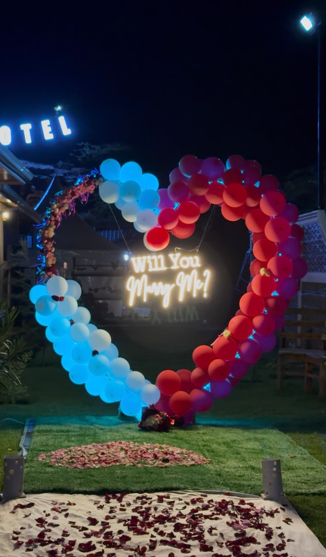 Heart-shaped balloon arch with a lit 'Will You Marry Me?' sign and rose petals in the Mawimbi Jiko garden at night
