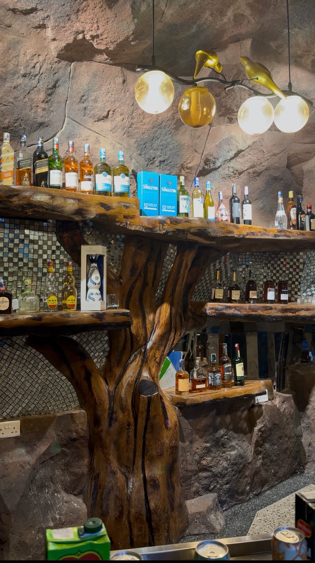 The rock alcove bar at Mawimbi Jiko with live-edge wooden shelving, mosaic backsplash and a bird-themed light fixture